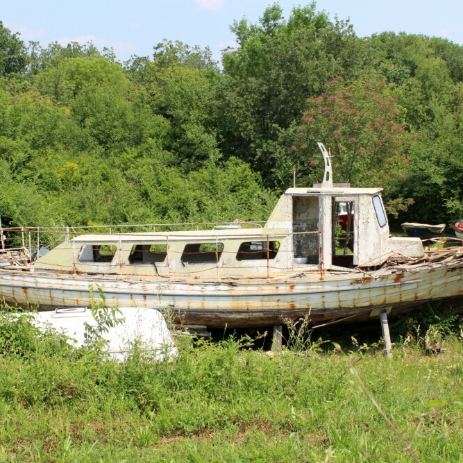 Abandoned old dilapidated wooden boat with broken windows, doors, wooden boards and rusted metal parts, left in backyard between uncut grass and forest vegetation on warm sunny day